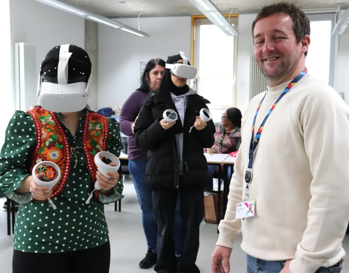 A smiling instructor stands in the classroom while a student wearing a VR headset and traditional attire engages in a virtual training session, with other students participating in the background. A smiling instructor stands in the classroom while a student wearing a VR headset and traditional attire engages in a virtual training session, with other students participating in the background.